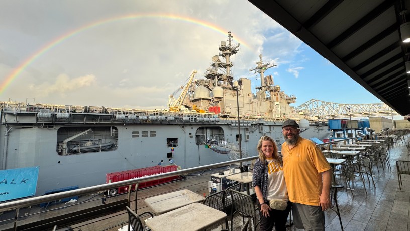 a man and woman stand in front of the USS Kearsarge under a rainbow